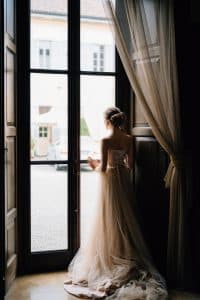 Bride stands at the large window of an old villa and looks into the courtyard. Lake Como. Back view