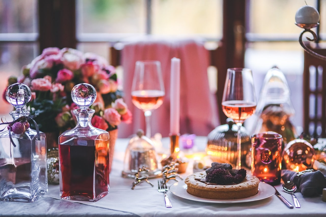 Eine in wohliges Licht getauchte Essenstafel mit gefüllten Gläsern und süßen Kuchen auf dem Teller mit einer warmen Deko mit rosa Akzenten